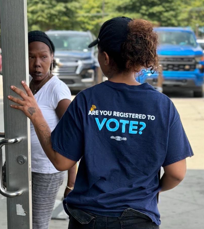 A New Engagement Fund canvasser at a doorway, with “ARE YOU REGISTERED TO VOTE?” on the back of the shirt